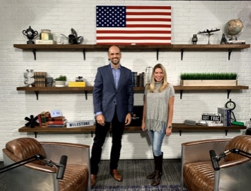 A picture of Gabriel Rio standing next to Leslie Beyer in front of a shelf with an American flag.